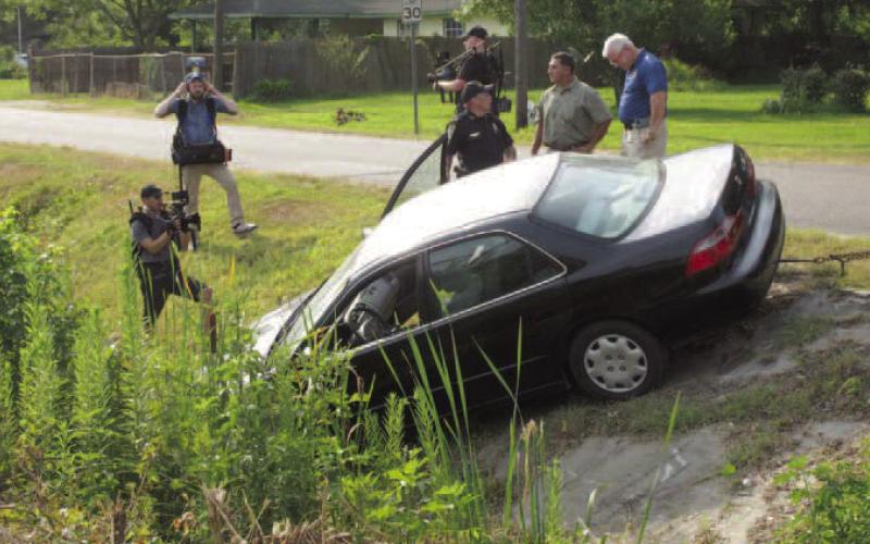 Vidor Police Chief Rod Carroll (blue shirt) is seen discussing the Kathy Page accident scene re-creation staged this week by the Vidor Police Department. With the chief are a Vidor Police Sergeant and the police department's accident reconstructionist. Surrounding the three men and the car are three unidentified civilians carrying video cameras who are photographing the scene. Archive Photo