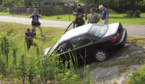 Vidor Police Chief Rod Carroll (blue shirt) is seen discussing the Kathy Page accident scene re-creation staged this week by the Vidor Police Department. With the chief are a Vidor Police Sergeant and the police department's accident reconstructionist. Surrounding the three men and the car are three unidentified civilians carrying video cameras who are photographing the scene. Archive Photo
