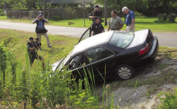 Vidor Police Chief Rod Carroll (blue shirt) is seen discussing the Kathy Page accident scene re-creation staged this week by the Vidor Police Department. With the chief are a Vidor Police Sergeant and the police department's accident reconstructionist. Surrounding the three men and the car are three unidentified civilians carrying video cameras who are photographing the scene. Archive Photo